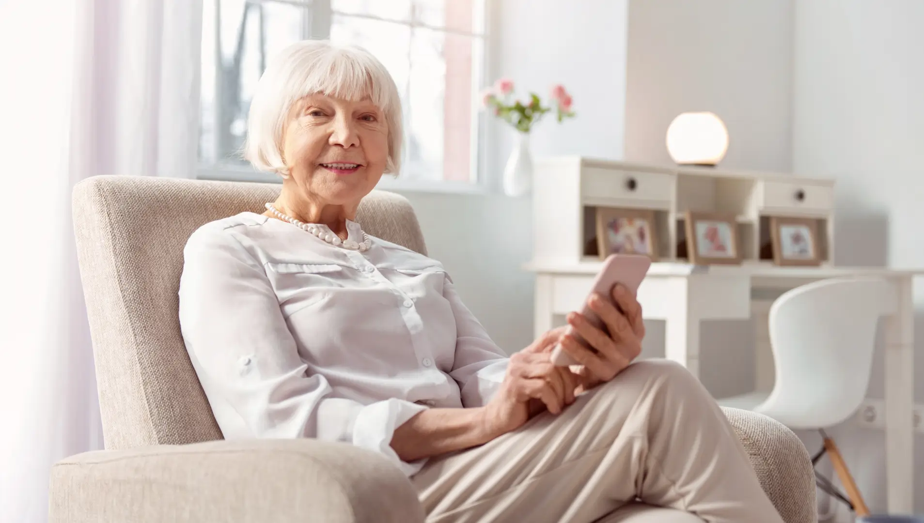 Eine ältere Dame sitzt in einem Sessel und tippt dabei auf einem Smartphone. Sie lächelt.