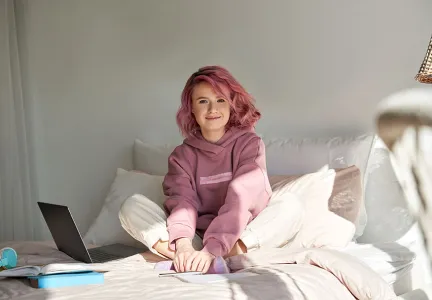 Auf dem Bild sitzt eine junge Frau mit rosa Haaren und einem rosa Hoodie lächelnd auf einem Bett. Neben ihr liegen ein Laptop und einige Bücher, was auf eine entspannte Lern- oder Arbeitsumgebung hinweist.