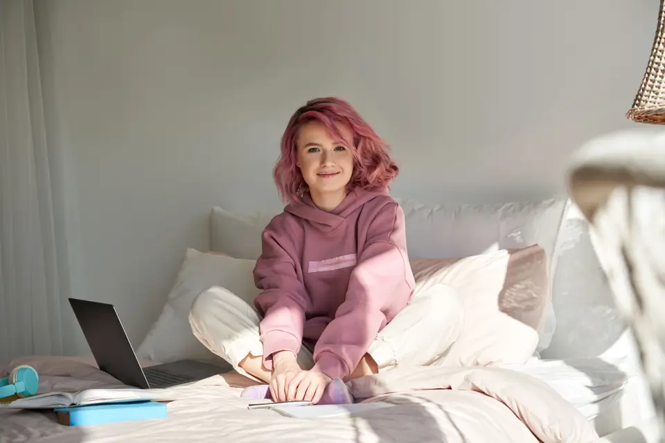 Auf dem Bild sitzt eine junge Frau mit rosa Haaren und einem rosa Hoodie lächelnd auf einem Bett. Neben ihr liegen ein Laptop und einige Bücher, was auf eine entspannte Lern- oder Arbeitsumgebung hinweist.