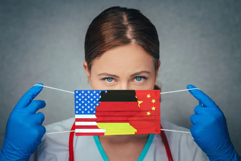 Das Bild zeigt eine Person, die blaue Einweghandschuhe trägt und eine Gesichtsmaske mit den Flaggen der USA, Deutschlands und Chinas hält.