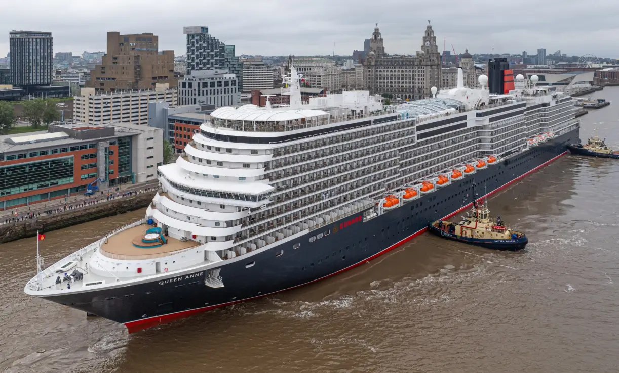 Das Kreuzfahrtschiff Queen Anne liegt, begleitet von Schleppern, vor den modernen und historischen Gebäuden von Liverpool.