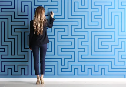 Auf dem Bild steht eine Frau mit langen, welligen Haaren in Geschäftskleidung vor einer großen blauen Wand, die mit einem Labyrinthmuster bedeckt ist. Sie scheint mit einem Stift auf die Wand zu zeichnen oder eine Lösung für das Labyrinth zu suchen.