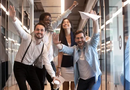 Auf dem Bild sind vier fröhliche Menschen in einem modernen Büro zu sehen, die gemeinsam Spaß haben und ein Papierflugzeug werfen. Sie lächeln und wirken ausgelassen und energiegeladen.