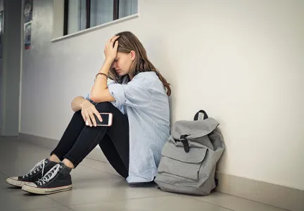 Auf dem Bild sitzt eine junge Frau mit einem traurigen Gesichtsausdruck auf dem Boden eines Flurs, während sie ein Smartphone in der Hand hält. Neben ihr steht ein grauer Rucksack.