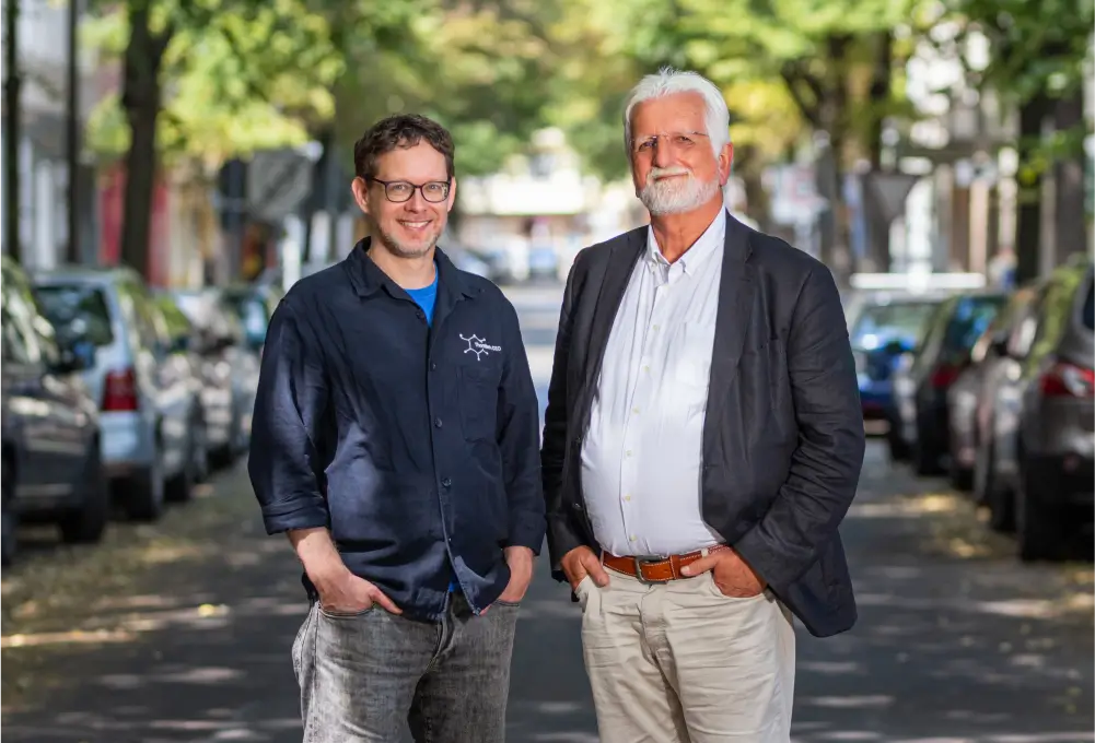 Zwei Männer stehen lächelnd auf einer von Bäumen gesäumten Straße. Der Mann links trägt eine Brille und ein dunkles Hemd, der Mann rechts hat graues Haar und trägt ein Jackett über einem weißen Hemd. Im Hintergrund sind parkende Autos und unscharfe Bäume zu sehen.