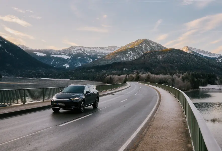 Ein grauer BYD Tang fährt über eine leicht gebogene Brücke, die einen ruhigen See in einer winterlichen Berglandschaft überquert. Die umliegenden Berge sind teilweise mit Schnee bedeckt und werden von sanftem, warmem Licht der untergehenden Sonne beleuchtet.