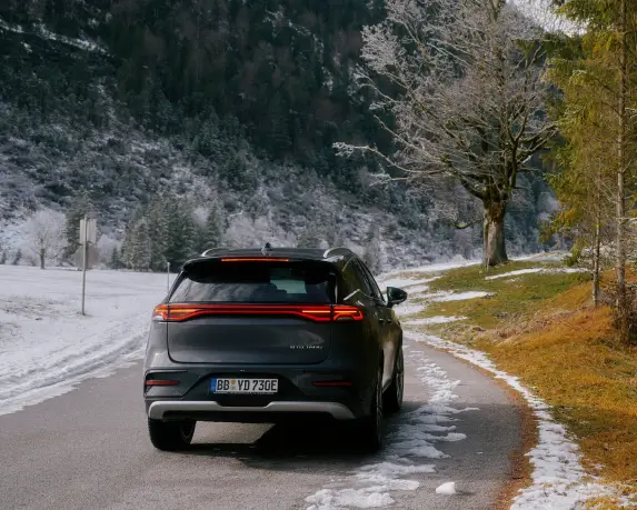 Ein grauer BYD Tang steht auf einer schmalen, teils verschneiten Straße in einer winterlichen Landschaft. Die Rückansicht des Autos zeigt die markanten Rücklichter, die eingeschaltet sind.