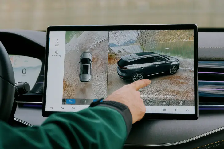 Das zentrale Touchscreen-Display im Innenraum eines BYD Tang zeigt die Kamerafunktion mit einer 360-Grad-Ansicht des Fahrzeugs. Links ist eine Vogelperspektive des Autos zu sehen, während rechts eine Seitenansicht angezeigt wird, die das Fahrzeug auf einem Schotterweg neben einem See zeigt.