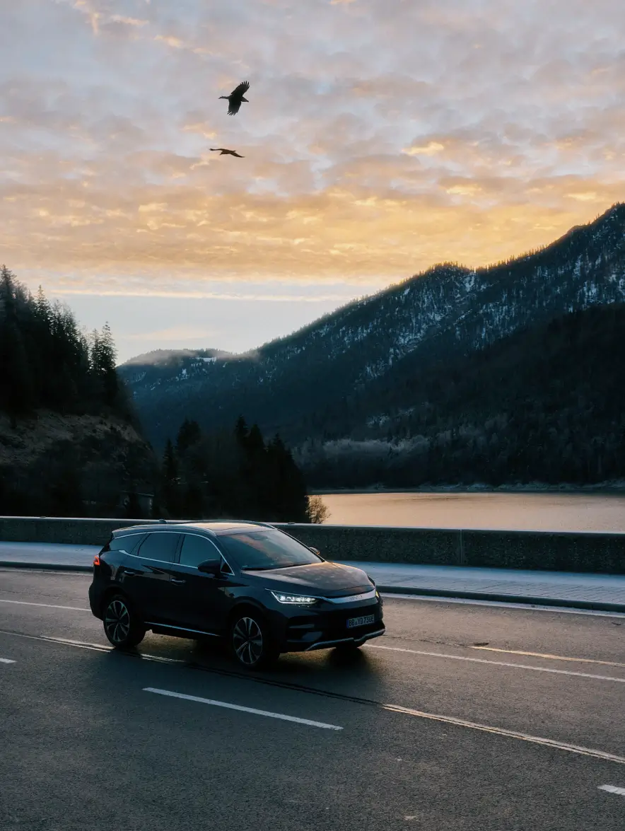 Ein grauer BYD Tang fährt auf einer asphaltierten Straße, die entlang eines Sees verläuft. Die Umgebung ist von bewaldeten Hügeln und Bergen umgeben, die mit Schnee bedeckt sind. Der Himmel leuchtet in warmen Gelb- und Orangetönen während des Sonnenuntergangs. Zwei Vögel fliegen über dem Auto am Himmel. Der See reflektiert das sanfte Licht des Himmels.