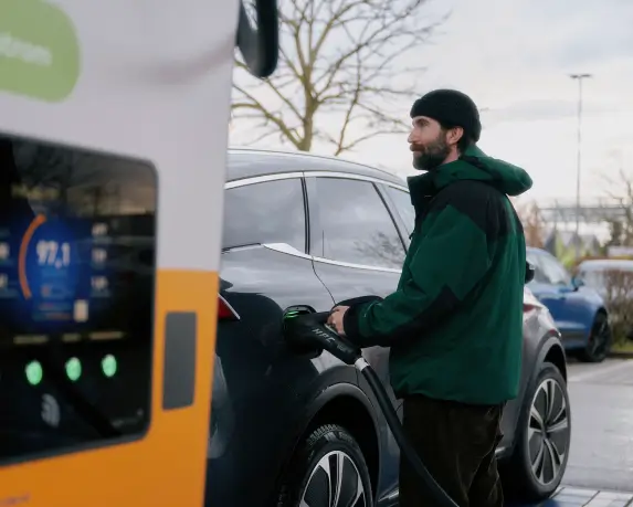 Ein Mann lädt einen grauen BYD Tang an einer Ladestation für Elektroautos auf. Er hält das Ladekabel, das mit dem Fahrzeug verbunden ist. Im Vordergrund ist unscharf die Anzeige der Ladestation zu sehen. Im Hintergrund befinden sich weitere Autos, kahle Bäume und ein bewölkter Himmel.