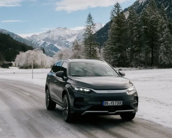 Ein grauer BYD Tang fährt auf einer schneebedeckten Straße durch eine winterliche Landschaft. Links und rechts der Straße sind schneebedeckte Felder und Bäume zu sehen, im Hintergrund erhebt sich eine beeindruckende Bergkulisse unter einem blauen Himmel mit vereinzelten Wolken. Das Fahrzeug ist von vorne sichtbar und die Scheinwerfer sind ausgeschaltet.