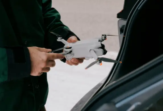 Nahaufnahme von Händen, die eine kleine weiße Drohne halten. Die Person steht neben einem offenen Kofferraum eines Autos, der teilweise im Bild zu sehen ist. Der Hintergrund zeigt eine asphaltierte Fläche und wirkt unscharf.