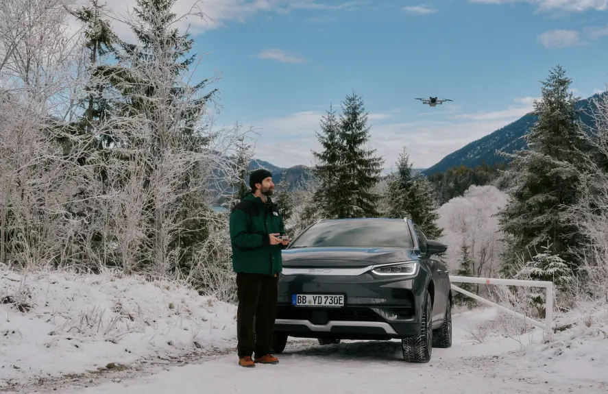 Ein Mann steht auf einem verschneiten Weg neben einem grauen SUV und steuert eine Drohne, die über ihm in der Luft schwebt. Die Umgebung ist von schneebedeckten Bäumen und einer winterlichen Berglandschaft geprägt. Der Himmel ist blau mit vereinzelten weißen Wolken.