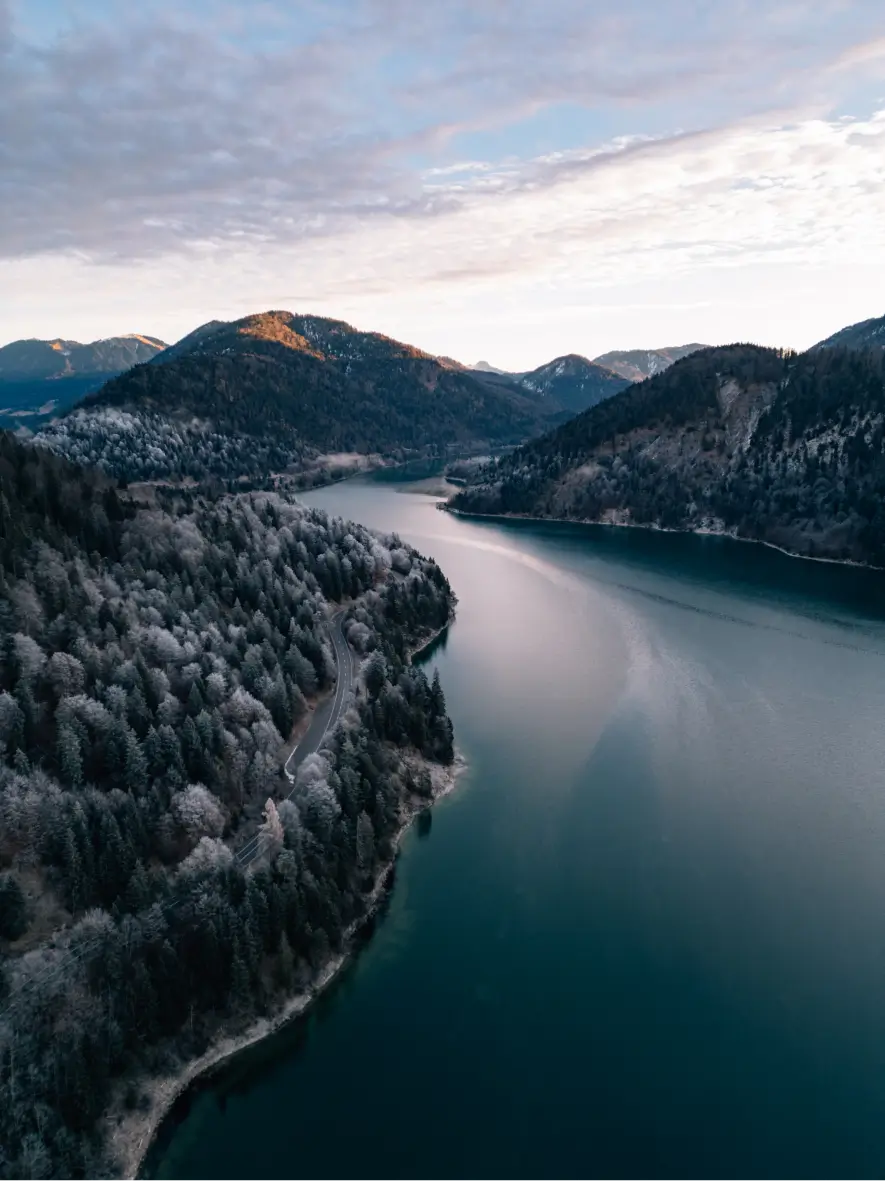 Luftaufnahme einer malerischen Landschaft mit einem geschwungenen, dunkelblauen See, der von bewaldeten Hügeln und Bergen umgeben ist. Die Bäume sind teils mit leichtem Raureif bedeckt. Eine Straße schlängelt sich entlang des Seeufers. Der Himmel ist bewölkt, mit vereinzelten Sonnenstrahlen, die die Bergspitzen beleuchten.