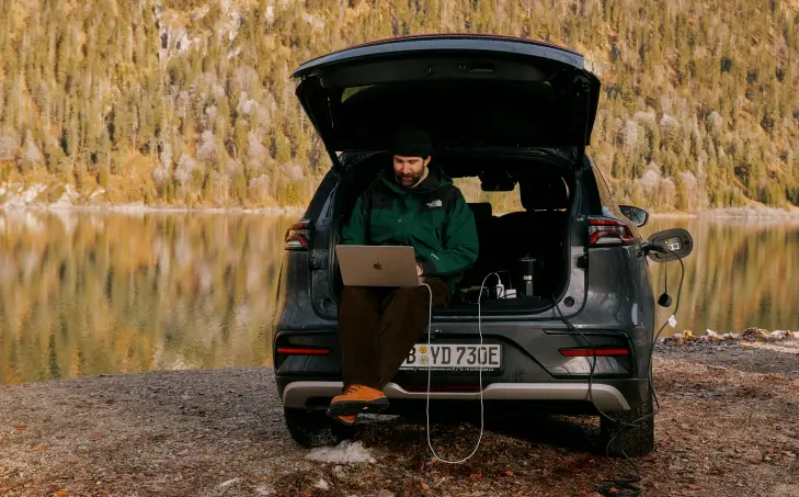 Eine Person sitzt im geöffneten Kofferraum eines Autos und arbeitet an einem Laptop, der über Kabel angeschlossen ist. Im Hintergrund ist ein ruhiger See zu sehen, umgeben von herbstlich gefärbtem Wald.