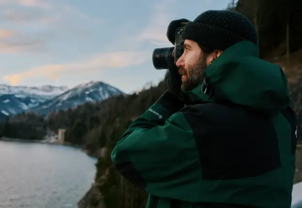 Eine Person mit dunkler Mütze und grüner Outdoor-Jacke fotografiert mit einer Kamera. Im Hintergrund sind ein See, bewaldete Hügel und schneebedeckte Berge unter einem leicht bewölkten Himmel zu sehen.