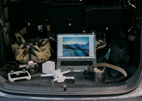 Geöffneter Kofferraum eines Autos, in dem verschiedene Gegenstände liegen. Zu sehen sind ein Laptop mit einem Landschaftsbild auf dem Bildschirm, eine Drohne, eine Fernsteuerung, Kameras, eine Tasche und weiteres Outdoor-Equipment. Die Szene vermittelt den Eindruck von Vorbereitung für Fotografie oder Outdoor-Aktivitäten.