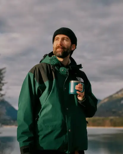 Eine Person mit dunkler Mütze und grüner Outdoor-Jacke steht vor einer Landschaft mit einem See und Bergen im Hintergrund. Die Person hält eine blaue Tasse in der Hand und blickt nach oben. Der Himmel ist teils bewölkt.