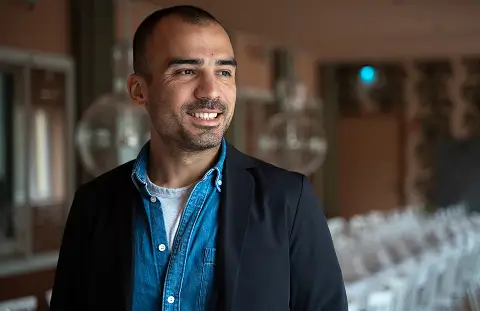 Eine Person in einem blauen Hemd und schwarzen Blazer steht in einem elegant eingerichteten Raum mit dekorativen Lampen und weiß gedeckten Tischen.