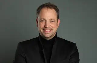 Eine Person in schwarzer Kleidung mit verschränkten Armen posiert selbstbewusst vor einem neutralen, grauen Hintergrund.