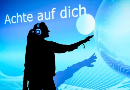 Eine Person mit Kopfhörern steht vor einem leuchtend blauen Hintergrund mit der Aufschrift „Achte auf dich“ und berührt scheinbar eine digitale Projektion mit ausgestrecktem Arm.