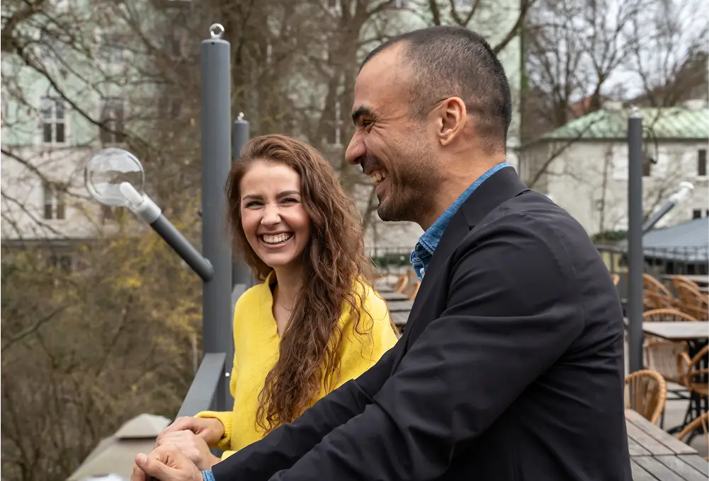 Zwei Personen stehen auf einer Terrasse mit modernem Geländer, umgeben von kahlen Bäumen und historischen Gebäuden im Hintergrund, während sie sich unterhalten.