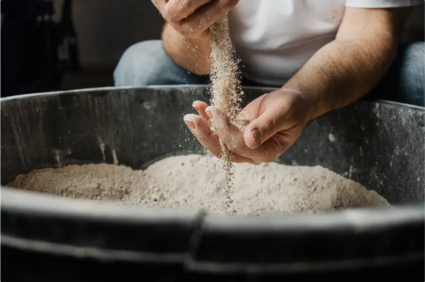 Hände lassen feinen Tonstaub in einen großen Behälter rieseln. Dieser Schritt verdeutlicht die sorgfältige Materialvorbereitung für die Produktion hochwertiger Keramiken.