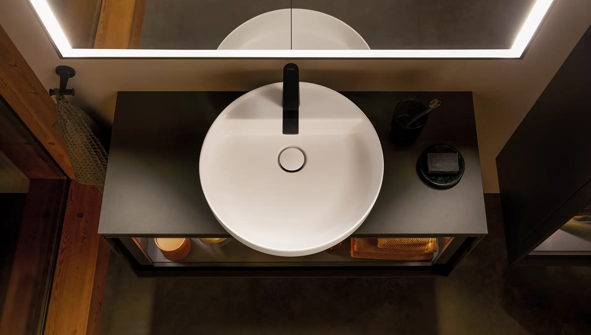 Ein rundes, weißes Waschbecken auf einer schwarzen Ablage mit minimalistischen Accessoires. Die Beleuchtung des Spiegels unterstreicht das elegante Design.