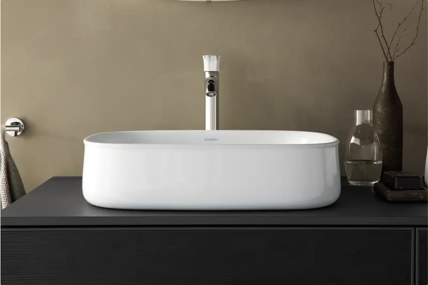 Weißes, ovales Waschbecken von Duravit auf einer dunklen Ablage. Dekorative Elemente wie eine Vase und ein Glas ergänzen das hochwertige Design.