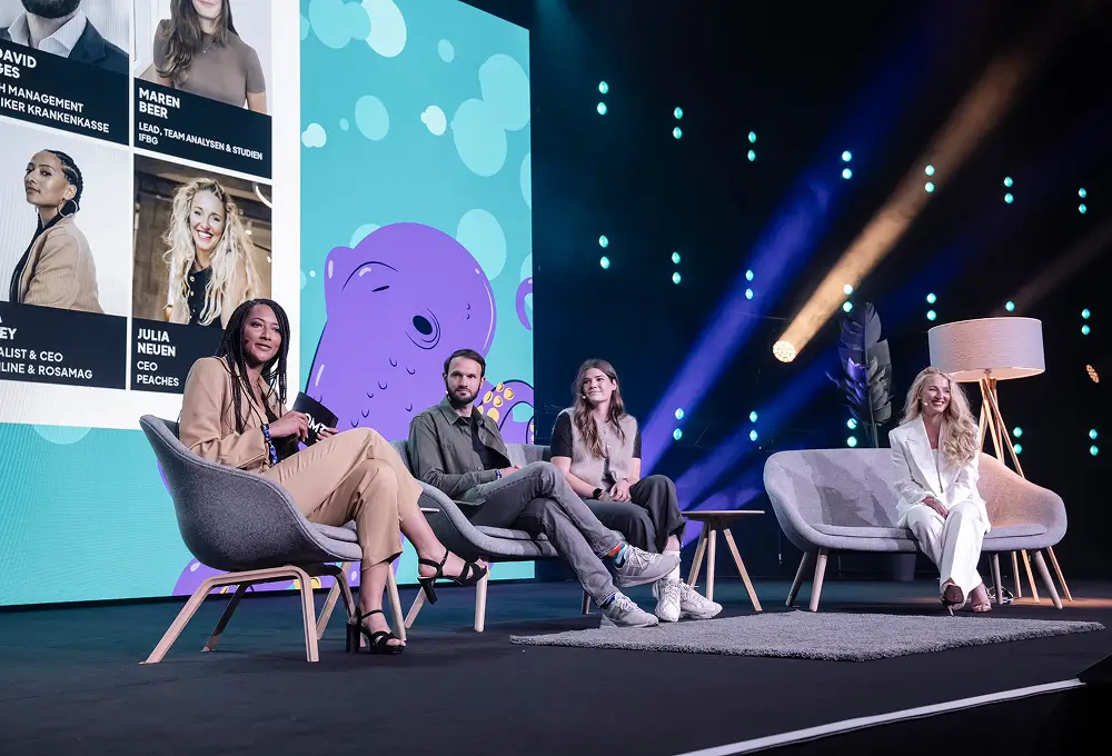 Eine Panel-Diskussion auf der Bühne der OMR 2025 mit vier Personen, die auf modernen Sesseln sitzen, vor einer bunten Leinwand mit einem Oktopus-Motiv.