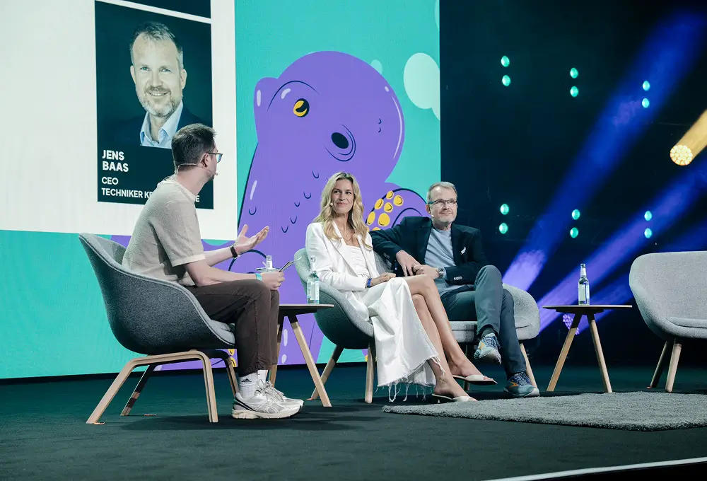 Drei Personen auf der Bühne diskutieren, während ein Moderator die Unterhaltung leitet; im Hintergrund ist die Leinwand mit einem Oktopus-Design zu sehen.