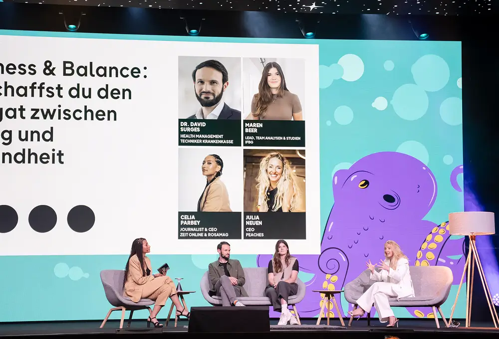 Eine Großaufnahme der Bühne mit vier Panel-Teilnehmern, die vor einer Leinwand mit dem Thema „Wellness & Balance“ sitzen.