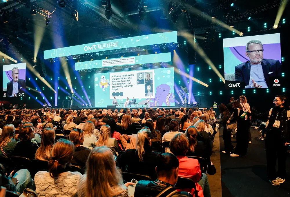 Die Blue Stage der OMR zeigt eine lebhafte Panel-Diskussion vor einem großen Publikum, mit einer farbenfrohen Leinwand und stimmungsvoller Beleuchtung im Hintergrund.