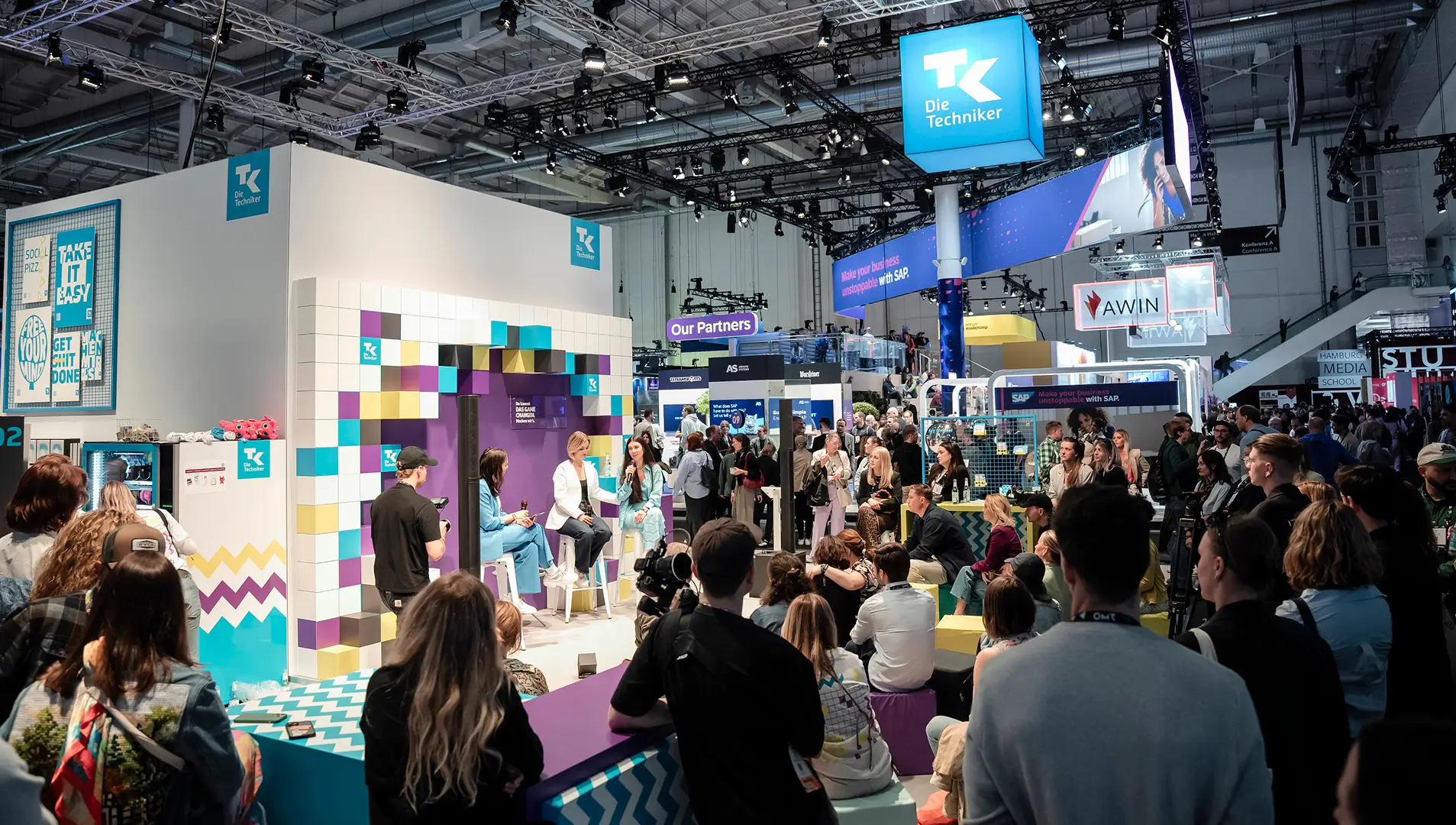 Der farbenfrohe Stand der Techniker Krankenkasse auf der Messe, umgeben von zahlreichen Besuchern und einer Live-Diskussion.