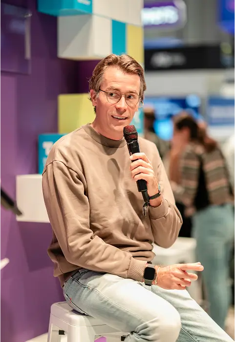Johannes Wimmer sitzt auf einem Hocker vor einer lila Wand und spricht in ein Mikrofon, während er gestikuliert.