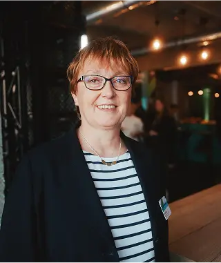 Eine Person in einem dunkelblauen Blazer und gestreiftem Oberteil steht vor einem modern gestalteten Hintergrund.