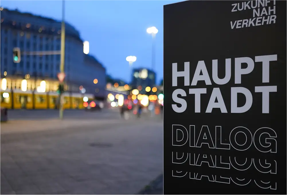 Ein Schild mit der Aufschrift „Hauptstadt Dialog“ im Vordergrund, während im Hintergrund eine belebte Straßenansicht mit Straßenbahn und Lichtern zu sehen ist.