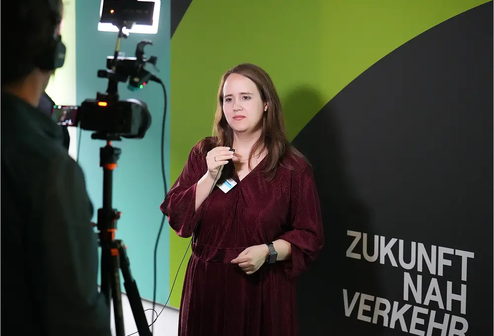 Eine Frau gibt ein Interview vor einer Kamera, stehend vor einer Wand mit der Aufschrift „Zukunft Nah Verkehr“.