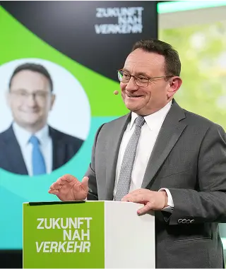 Ein Redner hält eine Präsentation an einem Pult mit der Aufschrift „Zukunft Nah Verkehr“ vor einer Leinwand mit grafischen Elementen.
