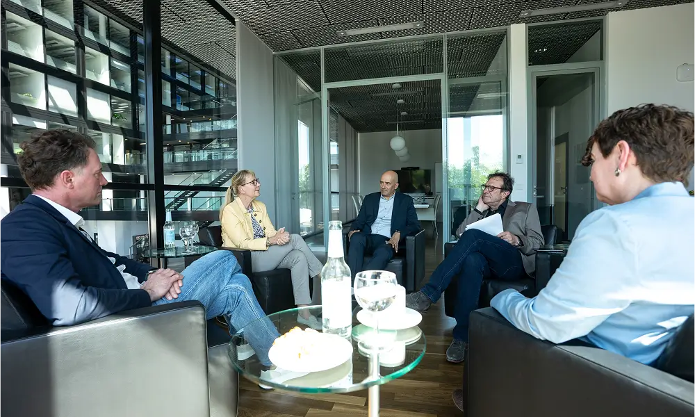 Fünf Personen sitzen in einem modernen, hellen Konferenzraum in einer Gesprächsrunde zusammen. Auf den Tischen stehen Getränke, Gläser und Snacks.