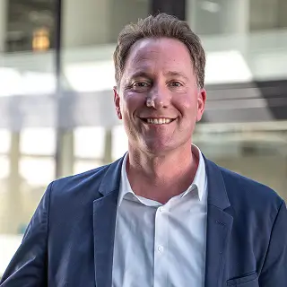 Dr. med. Holger Krönig steht in einem modernen, lichtdurchfluteten Gebäude und trägt ein weißes Hemd mit dunkelblauem Sakko.