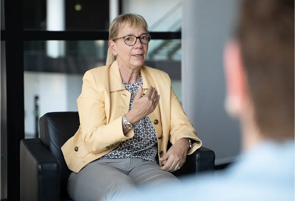 Eine Frau im gelben Blazer sitzt auf einem schwarzen Sessel und spricht gestikulierend mit einer weiteren Person im Vordergrund. Die Szene findet in einem modernen, hellen Innenraum statt.