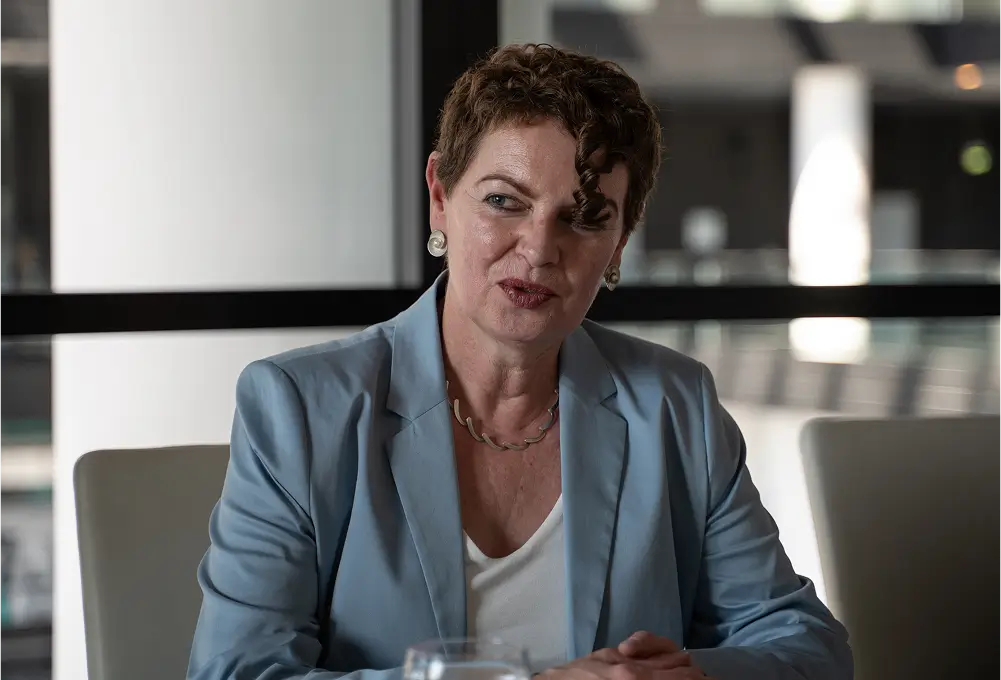 Eine Frau in einem hellblauen Blazer sitzt an einem Tisch in einem modernen, lichtdurchfluteten Raum. Vor ihr steht ein Glas