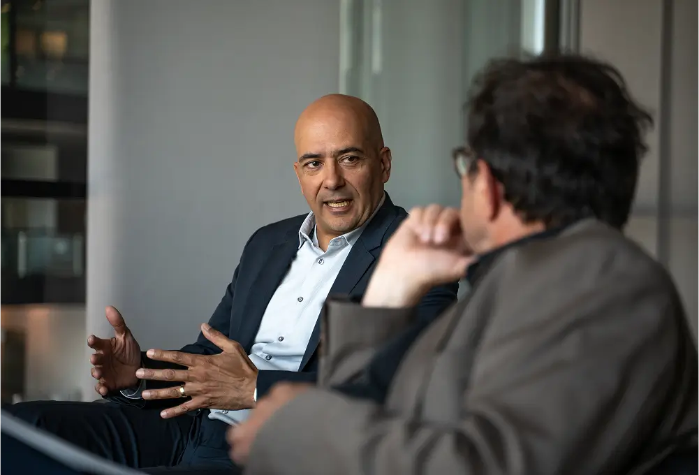 Ein Mann im Anzug sitzt in einem modernen Büro und spricht gestikulierend mit einer weiteren Person im Vordergrund.
