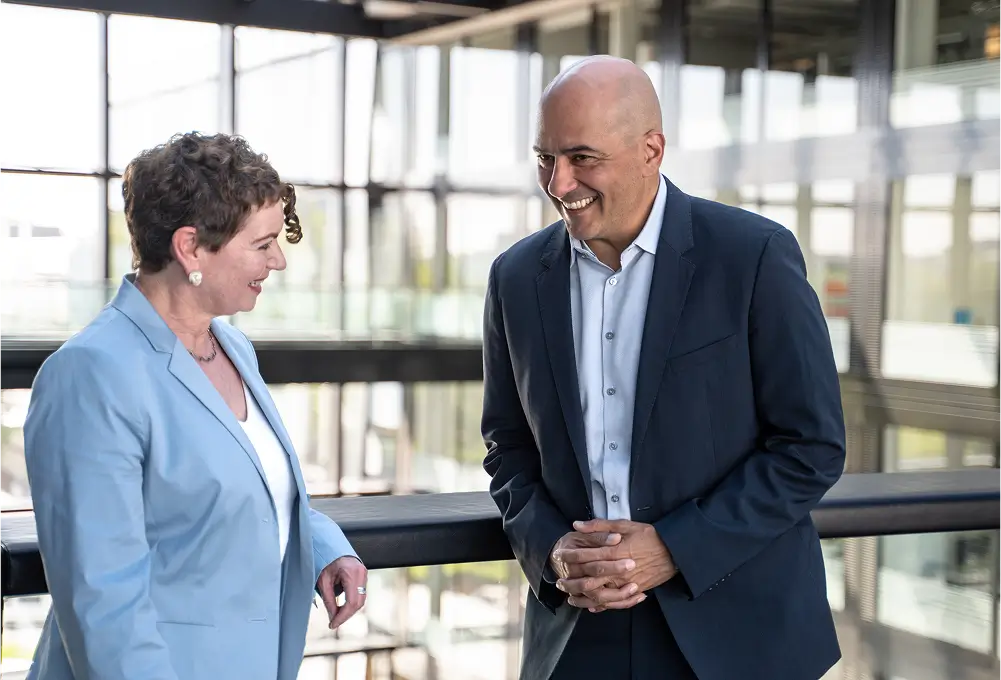 Zwei Personen im Business-Outfit stehen in einem modernen, lichtdurchfluteten Gebäude und unterhalten sich miteinander.
