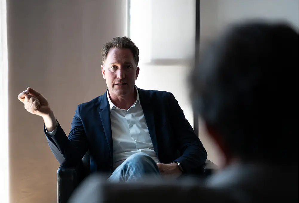 Ein Mann im dunklen Sakko sitzt entspannt auf einem Sessel und spricht mit einer weiteren Person im Vordergrund in einem modernen Raum.
