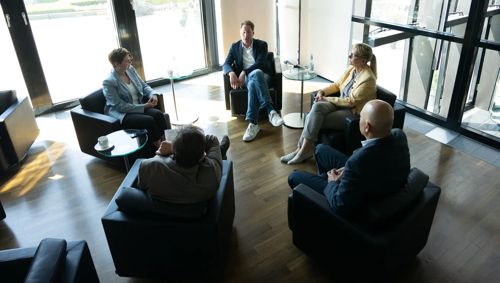 Vier Personen sitzen in einem modernen, lichtdurchfluteten Raum in Sesseln zu einem Gespräch im Kreis zusammen. Auf den kleinen Tischen stehen Getränke und Tassen.
