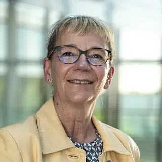 Susanne Busta steht in einem modernen, lichtdurchfluteten Gebäude und trägt einen gelben Blazer.