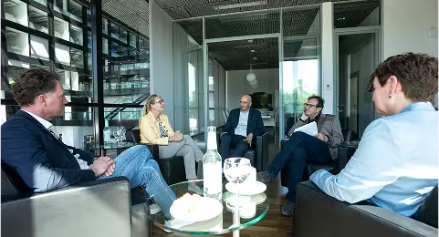 Fünf Personen sitzen in einem modernen, hellen Konferenzraum in einer Gesprächsrunde zusammen. Auf den Tischen stehen Getränke, Gläser und Snacks.