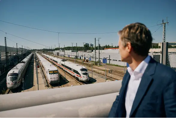 Mehrere ICE-Züge stehen nebeneinander auf Gleisen, im Vordergrund blickt eine Person von einer Brücke auf das Bahngelände.